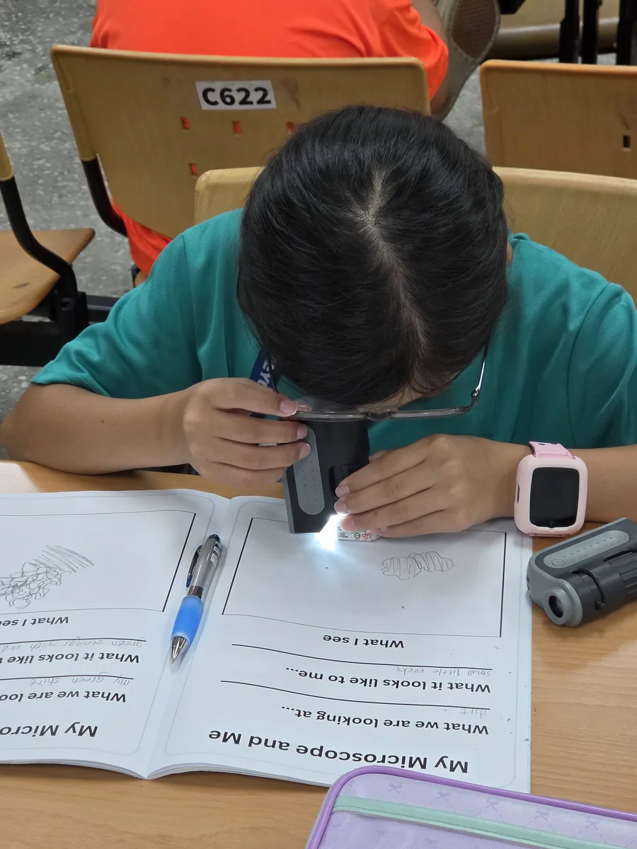 孩子低頭用手持顯微鏡看桌上的觀察手冊，手冊上印著 What I see 的畫圖格