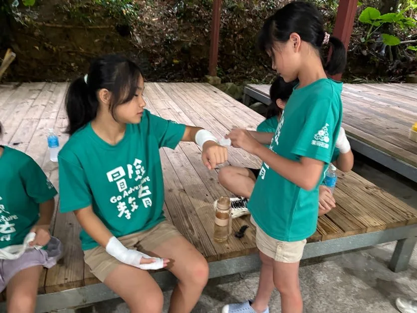 一個孩子坐在木板凳上，另一個孩子站在他面前，正在幫他手上的一處纏繃帶。