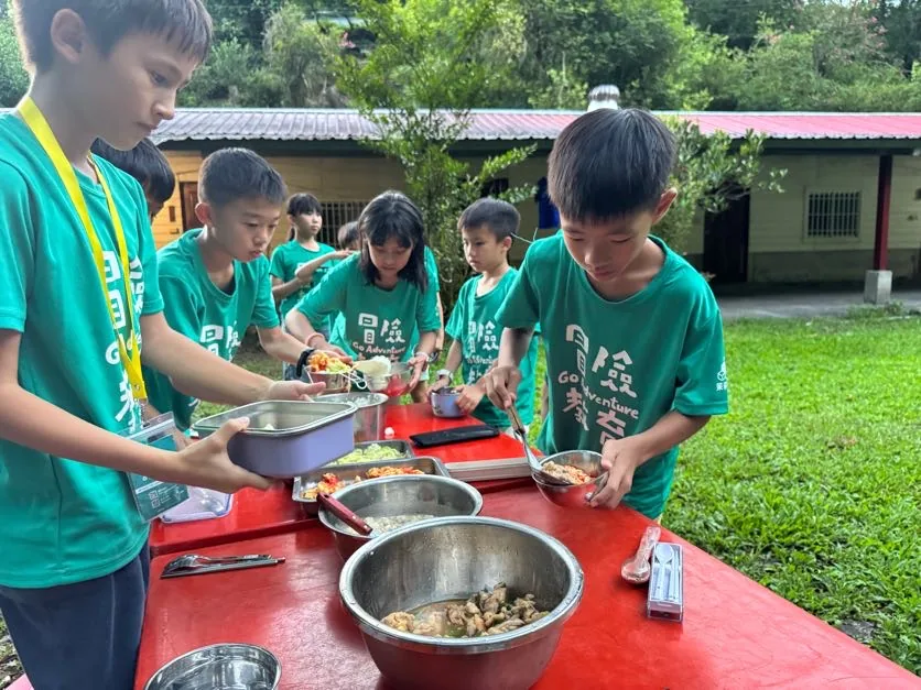 一群穿著同款 T-shirt 的孩子站在戶外餐桌前，用餐具從幾個不鏽鋼鍋盆裡分別盛取自己的一份。