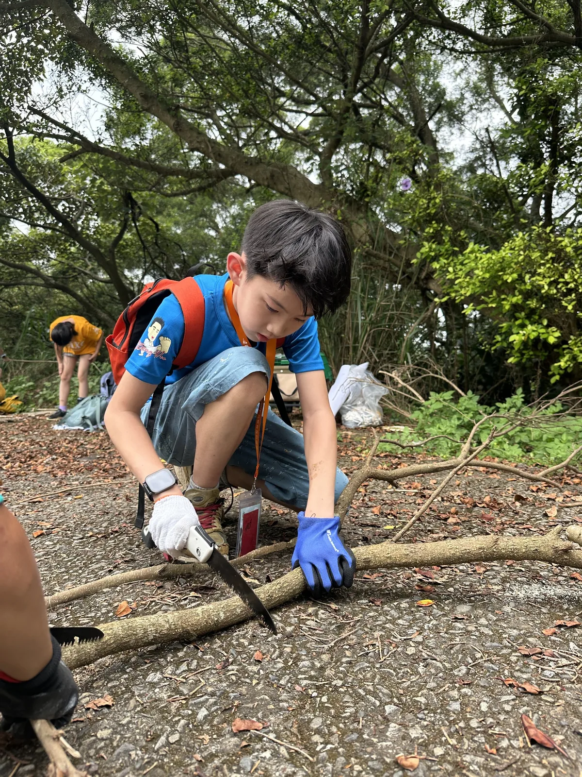 一個戴手套的孩子蹲在地上，用手鋸鋸一根樹枝。