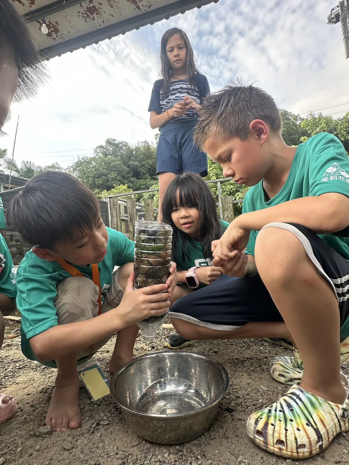 四個孩子圍蹲在地上，看著一個由寶特瓶裝著碎石與石塊做成的過濾裝置，水從瓶口滴進下方的不鏽鋼碗。