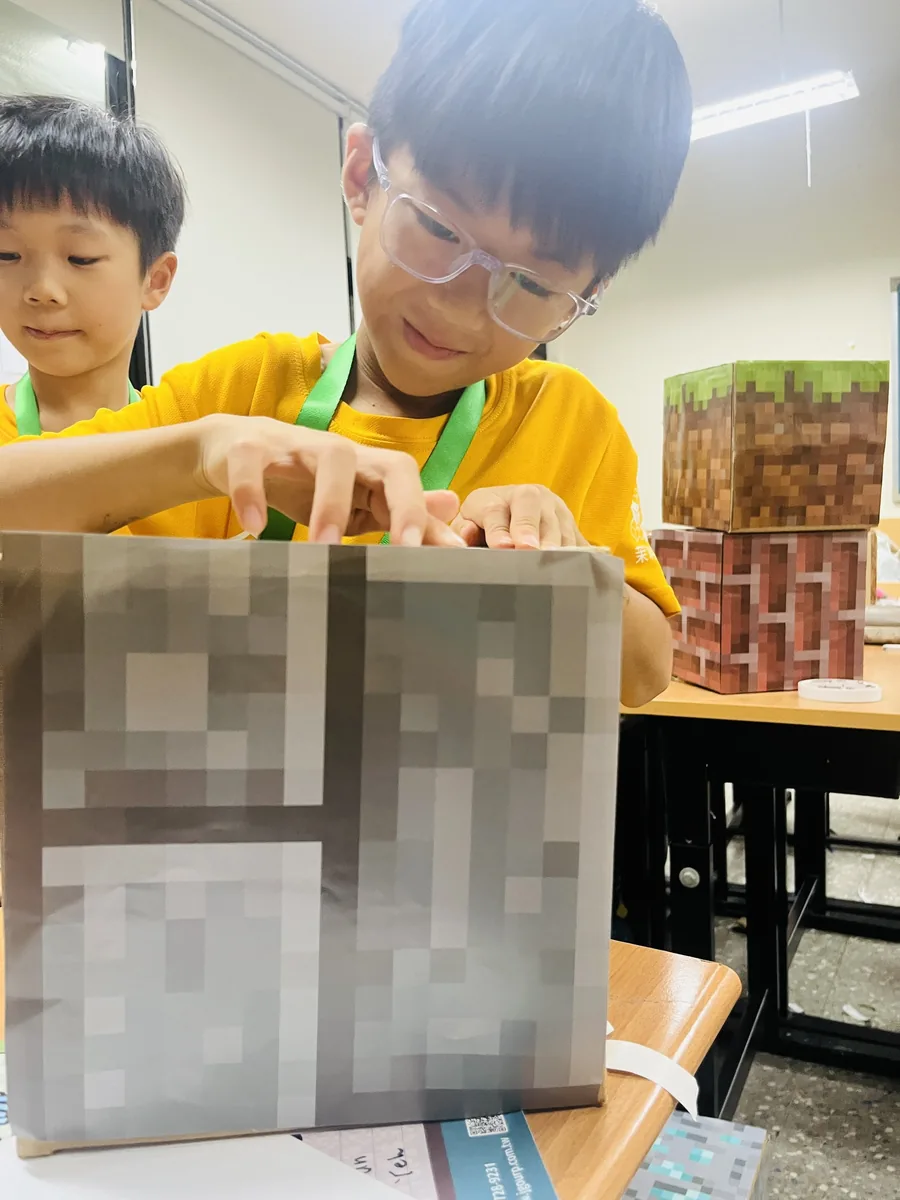 戴護目鏡的孩子製作像素方塊，桌上擺著草地方塊